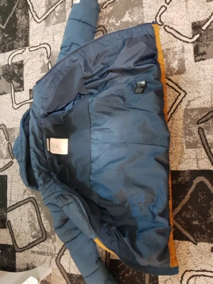 Blå och gul pufferjacka med huva - Säljer en snygg pufferjacka från Lindex i blått och gult. Jackan har huva, dragkedja och quiltad design som ger en cool look. Perfekt för kalla dagar och har en loose passform. Färgerna gör att du sticker ut på stan!