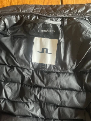 Svart dunväst från J.Lindeberg - Snygg svart dunväst från J.Lindeberg med quiltad design och hög krage. Västen har dragkedja framtill och två sidofickor med dragkedja. Perfekt för lager-på-lager och stilren look. Materialet är lätt och smidigt, vilket gör den enkel att bära.