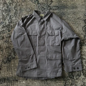 Grå overshirt från Tru-Spec - Snygg grå overshirt från Tru-Spec med fyra stora fickor framtill och klassisk krage. Tillverkad i slitstark blandning av polyester och bomull, perfekt för dig som gillar utility-vibe. Stängs med knappar vid ärmslut och har rak passform.