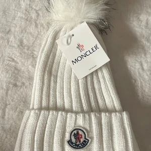 Vit Moncler mössa med pälsboll - Snygg ribbstickad vit mössa från Moncler med stor fluffig pälsboll på toppen. Mössan har Moncler-logga framtill och är tillverkad i mjukt stickat material. Perfekt för dig som vill ha en lyxig och trendig vinteraccessoar.