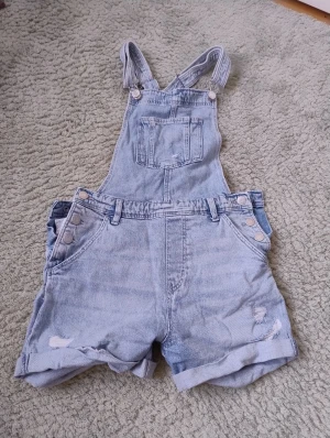 Ljusblå hängselshorts i denim - Säljer ett par ljusblå hängselshorts i denim. Klassisk jeanslook med bröstficka, knappar i sidorna och slitna detaljer framtill. Shortsen har upprullade benslut och flera fickor både fram och bak. Sparsamt använda.