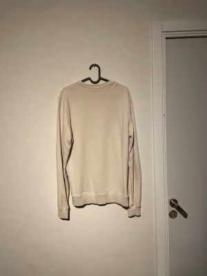 Beige sweatshirt från Lyle & Scott - Snygg beige sweatshirt från Lyle & Scott med klassisk rund hals och lång ärm. På bröstet sitter den ikoniska gula fågeln som broderad detalj. Tröjan har ribbade muddar och är gjord i mjuk bomull, perfekt för chill dagar.