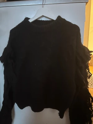 Svart stickad tröja med fransar - Cool svart stickad tröja med grova fransar längs ärmarna och ett flätat mönster framtill. Tröjan har rund hals och lång ärm, perfekt för dig som gillar statement-plagg med lite edge. Passar dig som vill ha en unik look och gillar att sticka ut. (större i storleken)