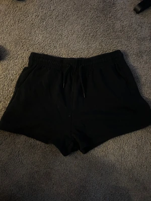 Svarta shorts med snörning - Svarta mjuka shorts med elastisk midja och snörning framtill. Klassisk och enkel design med fickor på sidorna, perfekta för chill eller träning. Materialet känns som bomull och passformen är loose för extra komfort.