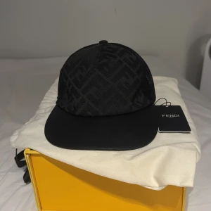 Svart Fendi keps med FF-mönster - Snygg svart keps från Fendi med ikoniskt FF-mönster i mesh och detaljer i guld på justerbar rem baktill. Skärmen är i svart skinn. Perfekt för dig som vill ha en exklusiv accessoar.har ej använt den nånting är ny skick  Skriv för mer frågor:)