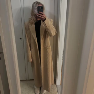 Beige lång kappa i manchester - Säljer en beige lång kappa i manchester med klassisk krage och knäppning framtill. Jackan är från NAKD i strl 36 och den går hela vägen ner på mig som är 168