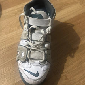 Nike Air More Uptempo vita sneakers - Nike Air More Uptempo sneakers i vitt med grå detaljer och stora AIR-bokstäver i mocka på sidorna. Skorna har snörning, perforerade paneler och en chunky platt sula med synlig Air-enhet. Snygg retrostil som sticker ut och passar perfekt till streetwear.