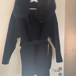 Svart kappa med pälsdetalj - Snygg svart kappa med bred krage i fluffig äkta päls och knytbälte i midjan. Jackan har långa ärmar och två stora fickor framtill. Perfekt för dig som vill ha en stilren och lyxig look under höst och vinter. Knappt använd, storlek S men passar M 
