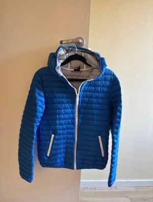 Blå pufferjacka från Colmar - Säljer en blå pufferjacka från Colmar med huva och vita dragkedjor. Jackan har quiltad design, två sidofickor med dragkedja och Colmar-logga på ärmen. Perfekt för dig som vill ha en snygg och sportig look under kyliga dagar. Storleken är 166cm 
