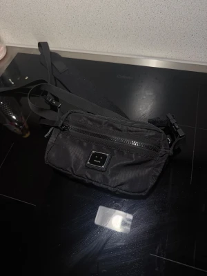 Svart axelväska från Acne Studios - Säljer en svart axelväska från Acne Studios med det klassiska smiley-emblemet framtill. Väskan har justerbar rem, dragkedja upptill och är tillverkad i slitstark syntet. Perfekt storlek för mobil, plånbok och nycklar.