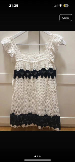 Vit spetsklänning med marinblå detaljer - Superfin kort vit klänning med spets och marinblå blommiga detaljer. Klänningen har breda axelband med volang och är lätt A-linjeformad. Perfekt för sommarens alla tillfällen och har en söt, romantisk vibe. Materialet är spets och klänningen är fodrad.