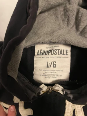 Vintage / Archive Hoodie - Svart hoodie från Aeropostale med stor vit broderad text 'AERO EST. 1987' på bröstet. Tröjan har huva med grått foder och vita snören, dragkedja framtill och två fickor. Materialet är 80% bomull. . Perfekt för vintern dagar.