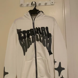 Vit hoodie Eternal Wraith med tryck - Säljer en vit hoodie från Eternal Wraith med svart grafiskt tryck på bröstet och ärmarna. Tröjan har huva med svart tryck och hel dragkedja framtill. Mjuk och skön polyester, perfekt för dig som gillar streetwear och unik design. Samt att den inte är använd alls 