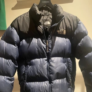 Blå och svart pufferjacka The North Face - Säljer en fet pufferjacka från The North Face i blått och svart. Jackan har hög krage, broderad logga på bröstet och ryggen, samt dragkedja framtill. Perfekt för kalla dagar. Storlek M.