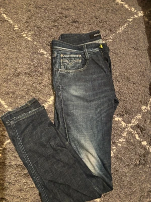 Replay Anbass jeans mörkblå slim fit - Säljer ett par Replay Anbass jeans i mörkblå tvätt med snygga slitningar och kontrastsömmar. Modellen är slim fit med klassisk femficksdesign och gul knappdetalj. Perfekt för dig som gillar stilrena jeans med lite edge. Storlek w33 L34