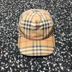 Burberry rutig keps i beige och svart - Snygg Burberry keps med klassiskt rutigt mönster i beige, svart och rött. Märket syns tydligt baktill med Burberry London England-etikett. Kepsen har böjd skärm och justerbar baksida. Materialet är bomull och polyester, perfekt för dig som vill ha en ikonisk streetstil.