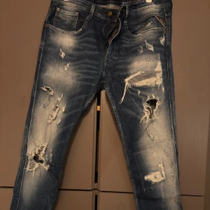 Replay Anbass blå slitna jeans 33/32 - Säljer ett par Replay Anbass jeans i blå tvätt med coola slitningar och trasiga detaljer framtill. Jeansen har klassisk femficksdesign, normal passform och är gjorda i jeansmaterial. Perfekt för dig som gillar en edgy streetstil.