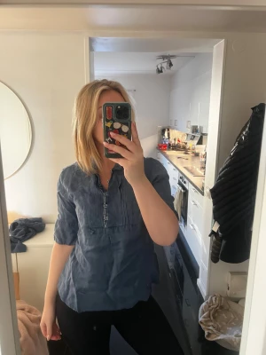 Mörkblå blus med knappar - Säljer en mörkblå blus med halvknäppning och dekorativa veck framtill. Blusen har korta ärmar och en avslappnad passform. Materialet känns lätt och luftigt, perfekt för dig som gillar stilrena och enkla plagg med en twist.