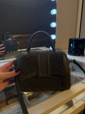 Svart handväska i päls från H&M - Snygg svart handväska från H&M med mjuk pälsstruktur och detaljer i skinnimitation. Väskan har ett stilrent handtag och avtagbar axelrem, perfekt för dig som gillar trendiga accessoarer. Insidan är fodrad och har en praktisk ficka.