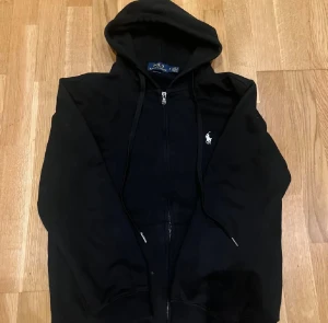 Svart hoodie från Polo Ralph Lauren - Svart hoodie från Polo Ralph Lauren med dragkedja och huva. Klassisk vit logga broderad på bröstet, snörning i huvan och praktiska fickor framtill. Tillverkad i mjuk bomull som är skön att ha på sig. Perfekt för chill dagar och streetwear. Passar S också 