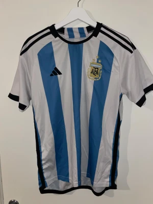 Argentina fotbollströja Adidas M - Säljer en officiell Argentina fotbollströja från Adidas i storlek M. Tröjan har klassiska blå och vita vertikala ränder, svarta detaljer vid krage och ärmslut samt AFA-emblem med tre stjärnor på bröstet. Tillverkad i lätt och ventilerande polyester.