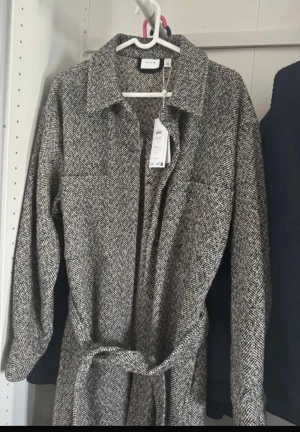 Grå långklänning från VILA med bälte - Säljer en långklänning från VILA i gråmelerat fiskbensmönster. Klänningen har lång ärm, klassisk krage och knytskärp i midjan för en snygg siluett. Perfekt för dig som gillar stilrena detaljer och vill ha en dressad look.