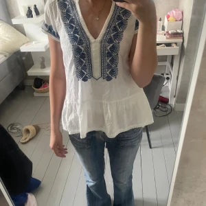 Vit blus med blå broderi Odd Molly - (Priset kan diskuteras) Supersöt vit blus från Odd Molly med blå broderade detaljer längs v-ringningen och på ärmarna. Blusen har korta ärmar, volang nedtill och är gjord i mjuk bomull. Perfekt för dig som gillar boho-vibe och snygga broderier.