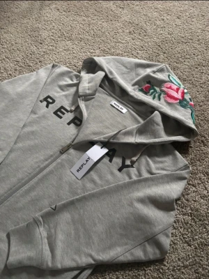 Grå hoodie med broderad ros från Replay - Grå hoodie från Replay med dragkedja och huva. Snygg broderad ros i rosa och grönt på huvan, samt svart Replay-tryck på bröstet. Mjuk bomullskvalitet och ribbade muddar vid ärmslut och nederkant. Perfekt för chill dagar. Fett fräsch priset ej hugget i sten