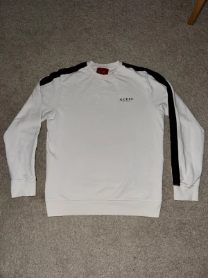 Vit sweatshirt från Guess med svarta detaljer - Vit långärmad sweatshirt från Guess med svarta ränder längs axlar och ärmar. Tryckt Guess-logga på bröstet. Mjuk och skön bomullskvalitet, perfekt för chill dagar. Klassisk passform med ribbade muddar vid ärmslut och nederkant.