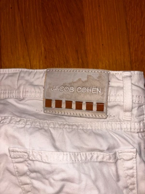 Vita Jacob Cohën byxor, storlek 32 - Snygga vita byxor från Jacob Cohën med klassisk passform och coolt mönstrat innerfoder. Byxorna har raka ben, fem fickor och exklusiva detaljer som märkespatch i mocka med bruna rutor. Perfekt för dig som gillar stilrena och lyxiga plagg.