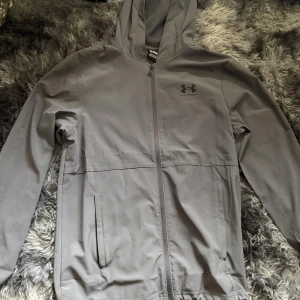 Grå vindjacka från Under Armour - Snygg grå vindjacka från Under Armour med huva och hel dragkedja framtill. Jackan har två sidofickor och diskret logga på bröstet. Perfekt för träning eller chill dagar ute. Långärmad och med stilren design som passar till det mesta.
