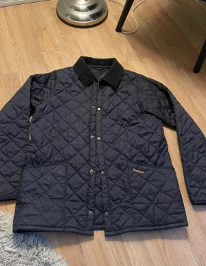 Svart quiltad Barbour jacka XXL (Barnstorlek) - Quiltad svart jacka från Barbour i storlek (barn XXL). Klassisk design med krage i kontrastmaterial, tryckknappar framtill och två stora fickor. Jackan har Barbour-logga broderad på fickan och är perfekt för dig som gillar stilren och tidlös look.