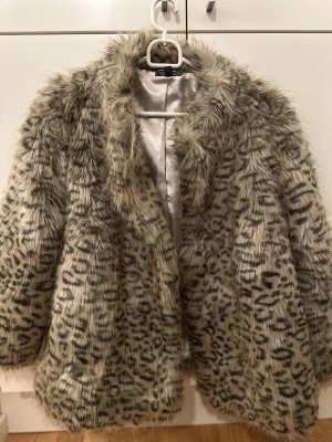 Topshop leopardmönstrad jacka, storlek 38 - Säljer en fluffig och snygg pälsjacka från Topshop med leopardmönster i beige och svart. Jackan har en bred krage, långärmade ärmar och är tillverkad i syntetpäls med glansigt innerfoder. Perfekt för dig som vill sticka ut och hålla dig varm.