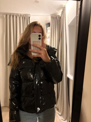 Svart pufferjacka Calvin Klein Jeans - Säljer en svart, glansig pufferjacka från Calvin Klein Jeans med huva och dragkedja. Jackan har CK-logga på bröstet och Calvin Klein Jeans-brodyr på ärmen. 