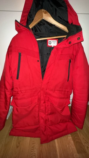 Röd parkajacka från Jack & Jones M - Parkajacka i stark röd färg från Jack & Jones Core, med huva och svarta dragkedjor på bröstet. Jackan har stora fickor framtill och är fodrad med svart innerfoder. Perfekt för kalla dagar och har en clean, urban look.
