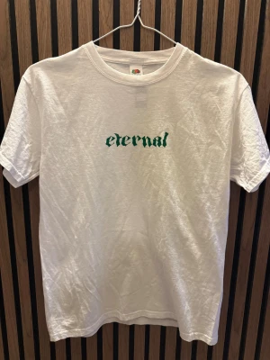 Vit t-shirt Eternal logo - Vit t-shirt från Fruit of the Loom i storlek S med trycket 'eternal' i grönt på bröstet. Klassisk rund hals och korta ärmar. Tillverkad i mjuk bomull som känns skön mot huden. Perfekt för dig som gillar enkel och stilren streetwear.