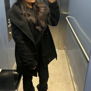 Svart kappa med bälte och huva - Säljer en svart kappa med stor huva och bälte i midjan. Jackan har knappar framtill och en snygg, lite oversized passform. Perfekt för dig som gillar stilrena och enkla plagg med lite edge. Materialet känns mjukt och värmande, passar bra till höst och vinter.