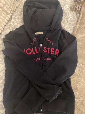 Marinblå zip up hoodie från Hollister - Marinblå zip up hoodie från Hollister med rosa tryck på bröstet och texten 'California Surf Club'. Tröjan har huva, långa ärmar och dragkedja framtill. Perfekt för chill dagar och har klassisk streetstil.
