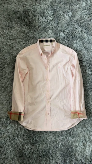 Ljusrosa Burberry skjorta - Ljusrosa skjorta från Burberry med klassiska rutiga detaljer på insidan av kragen och ärmsluten. Skjortan har knappar framtill, lång ärm och broderad logga på bröstet. Tillverkad i mjuk bomull med lite stretch för extra komfort.