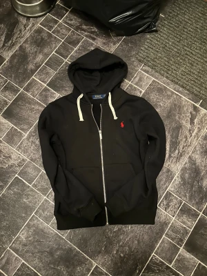 Svart hoodie från Polo Ralph Lauren - Svart hoodie från Polo Ralph Lauren med vit snörning och dragkedja framtill. Klassisk röd broderad logga på bröstet och huva. Tröjan har långa ärmar och är gjord i mjuk bomull, perfekt för chill dagar. Snygg och enkel att matcha med jeans eller joggers.