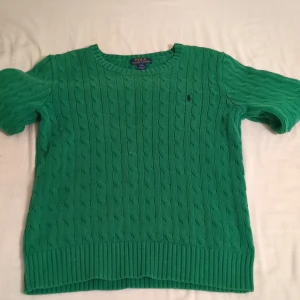 Grön stickad tröja Polo Ralph Lauren - Snygg grön stickad tröja från Polo Ralph Lauren med klassiskt kabelstickat mönster och ribbade muddar. Tröjan har rund hals och korta ärmar, perfekt för dig som gillar clean still och vill ha något som sticker ut lite extra.