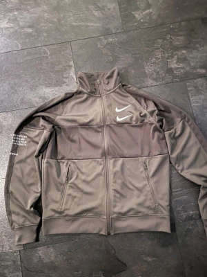 Grå/grön  Nike vindjacka med dragkedja - Snygg grå vindjacka från Nike med två vita broderade swoosh-loggor på bröstet. Jackan har hel dragkedja, höga krage och två sidofickor med dragkedja. Materialet är mjukt och lätt polyester, perfekt för sportiga dagar. Tryckt text på ena ärmen och ryggen. Den är i väldigt fint skick och har ett fantastiskt pris. Kontakta vid frågor.