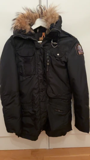 Svart Parajumpers vinterjacka med päls - Svart Parajumpers vinterjacka i slim fit med snygg päls på huvan, flera fickor och ikonisk patch på ärmen. Jackan har dragkedja, knappar och coola gula detaljer. Perfekt för kalla dagar när du vill hålla stilen. Pris kan även diskuteras. Trasig dragkedja men det går att fixa  