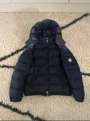 Mörkblå dunjacka från Moncler - Snygg mörkblå dunjacka från Moncler med huva och dragkedja framtill. Jackan har puffad design, två sidofickor med dragkedja och Moncler-logga på ärmen. Materialet är polyester och nylon med äkta dunfyllning. Perfekt för kalla dagar och riktigt stilren look.
