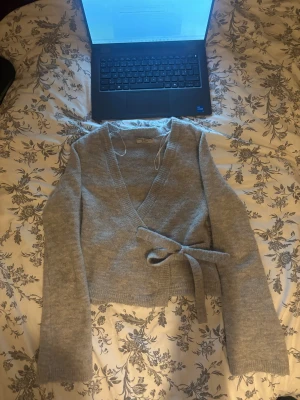 Grå omlottkofta från Gina Tricot - Supermysig grå omlottkofta från Gina Tricot i stickat material. Koftan har långa ärmar, v-ringad hals och ett snyggt knytband i midjan. Perfekt för att få en chill och stilren look, passar till jeans eller kjol. Premium Quality.