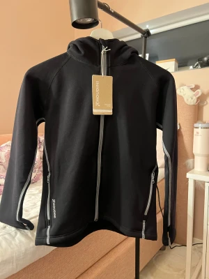 Svart Houdini fleece med huva - HELT NY OCH OANVÄND MED LAPPEN KVAR!! Svart vindjacka från Houdini med huva och grå detaljer längs dragkedjorna. Jackan har hel dragkedja framtill, två sidofickor med dragkedja och diskret Houdini-logga på bröstet och huvan. Perfekt för utomhusaktiviteter och chill dagar.