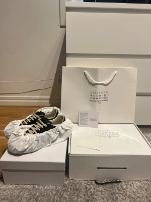 Maison Margiela svarta sneakers - Maison Margiela sneakers i svart skinn och mocka med beige snörning och gummisula i brun ton. Klassisk rund tå och platt sula, med diskret branding under sulan. Snygg minimalistisk design som passar till streetwear och trendiga outfits. 