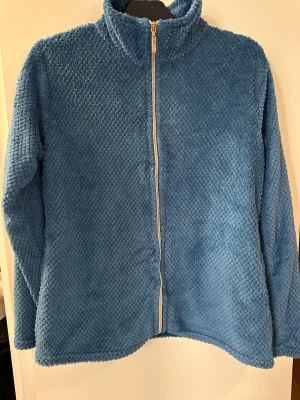 Blå teddyjacka med dragkedja strl: S - Mjuk och fluffig blå teddyjacka med hel dragkedja framtill och hög krage. Jackan är tillverkad i 100% polyester och har långärmade ärmar. Perfekt för dig som gillar chill och comfy vibes under kyliga dagar.