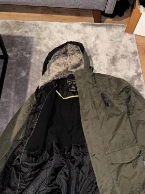 Vintage jack grön med päls - Parkajacka i militärgrön färg från Urban Mind, med broderad text på ärmen och flera fickor framtill. Jackan har huva med fuskpäls och quiltat svart innerfoder. Perfekt för kalla dagar med sin långa passform och praktiska detaljer.