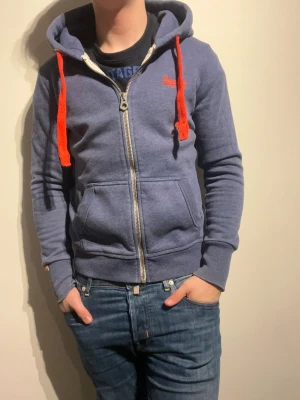 Superdry Zip Hoodie - Hej! Säljer nu denna väldigt snygga Superdry Zip Hoodien.  Storlek M Medium   Färg: Blå och orange  Skick: Gott  – i fint använt skick   Skickas spårbart eller hämtas upp efter överenskommelse. Vid intresse – hör gärna av dig.  Skriv vid frågor eller ifall du vill ha fler bilder:) 